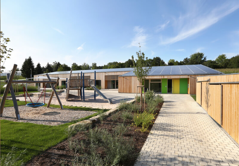 holzfarbene Kita Gebäude mit Rasenfläche, Schaukel und Rutsche